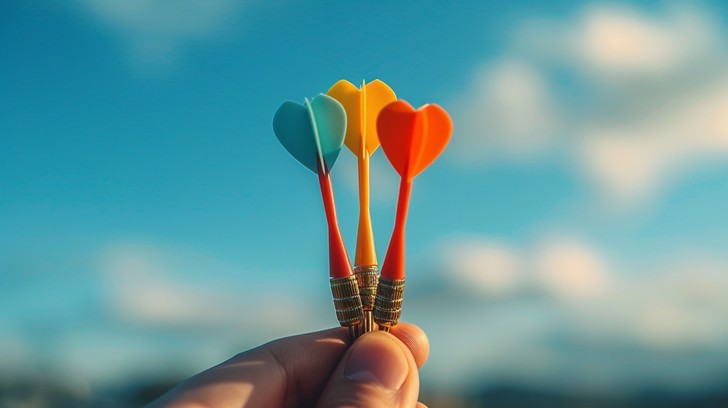 A hand holds three colorful tungsten darts with heart-shaped tails against a beautiful blue sky backdrop.